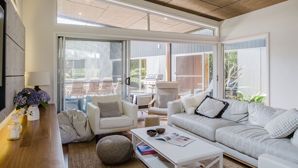 A modern living room with a white sofa, a coffee table, and a view of an outdoor patio with chairs and a grill. - The Magic Beach House (Narrawallee)