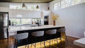 A modern kitchen with a large island, bar stools, stainless steel appliances, and large windows. - The Magic Beach House (Narrawallee)