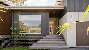 A modern house with a large window, a brick wall, and a set of stairs leading to the entrance. - The Magic Beach House (Narrawallee)