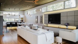 A modern living room with a large white sofa, a dining area with a white table and chairs, and a kitchen area with a bar counter. - The Magic Beach House (Narrawallee)