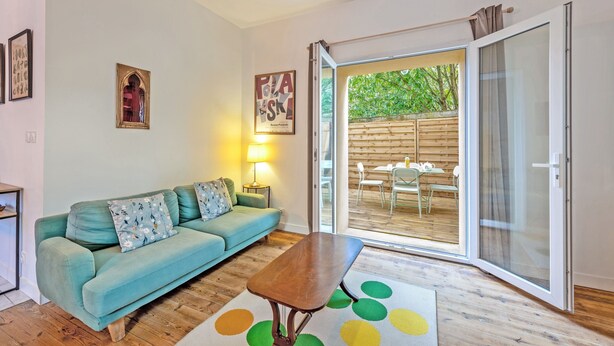 A living room with a teal sofa, a wooden coffee table, and a glass door leading to an outdoor area with a wooden deck and chairs. - Le 52 - Proche du GR34 (Morlaix)