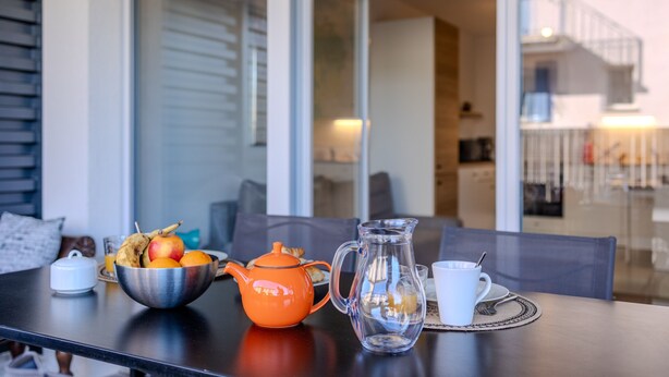A dining table with a metal bowl of fruit, an orange teapot, a clear glass pitcher, and a white cup on a placemat. - Casa Kenez - A 5min de la plage (Cavalaire-sur-Mer)
