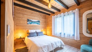 A cozy wooden cabin bedroom with a bed, a nightstand, a window with curtains, and a painting on the wall.