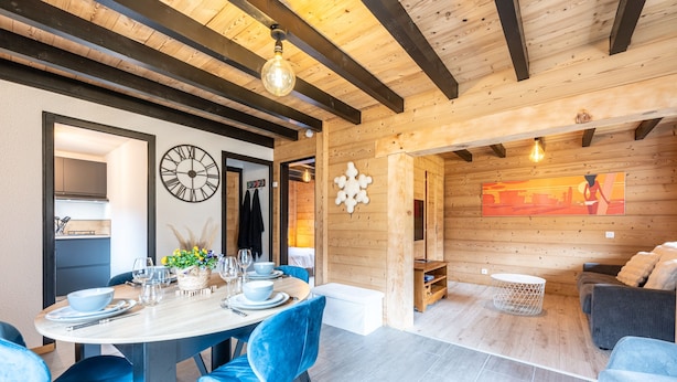 A modern interior with a wooden ceiling, a dining area with blue chairs, a round table, and a sofa in the background.