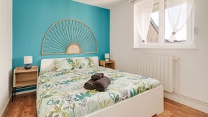 A bedroom with a teal accent wall, a bed with a leaf-patterned bedspread, a wooden headboard, and a window with sheer curtains. - Aux 4 saisons - Proche de Gérardmer (Corcieux)