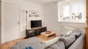 A living room with a grey sofa, a wooden coffee table, a TV on a black stand, and a window with sheer curtains. - Aux 4 saisons - Proche de Gérardmer (Corcieux)
