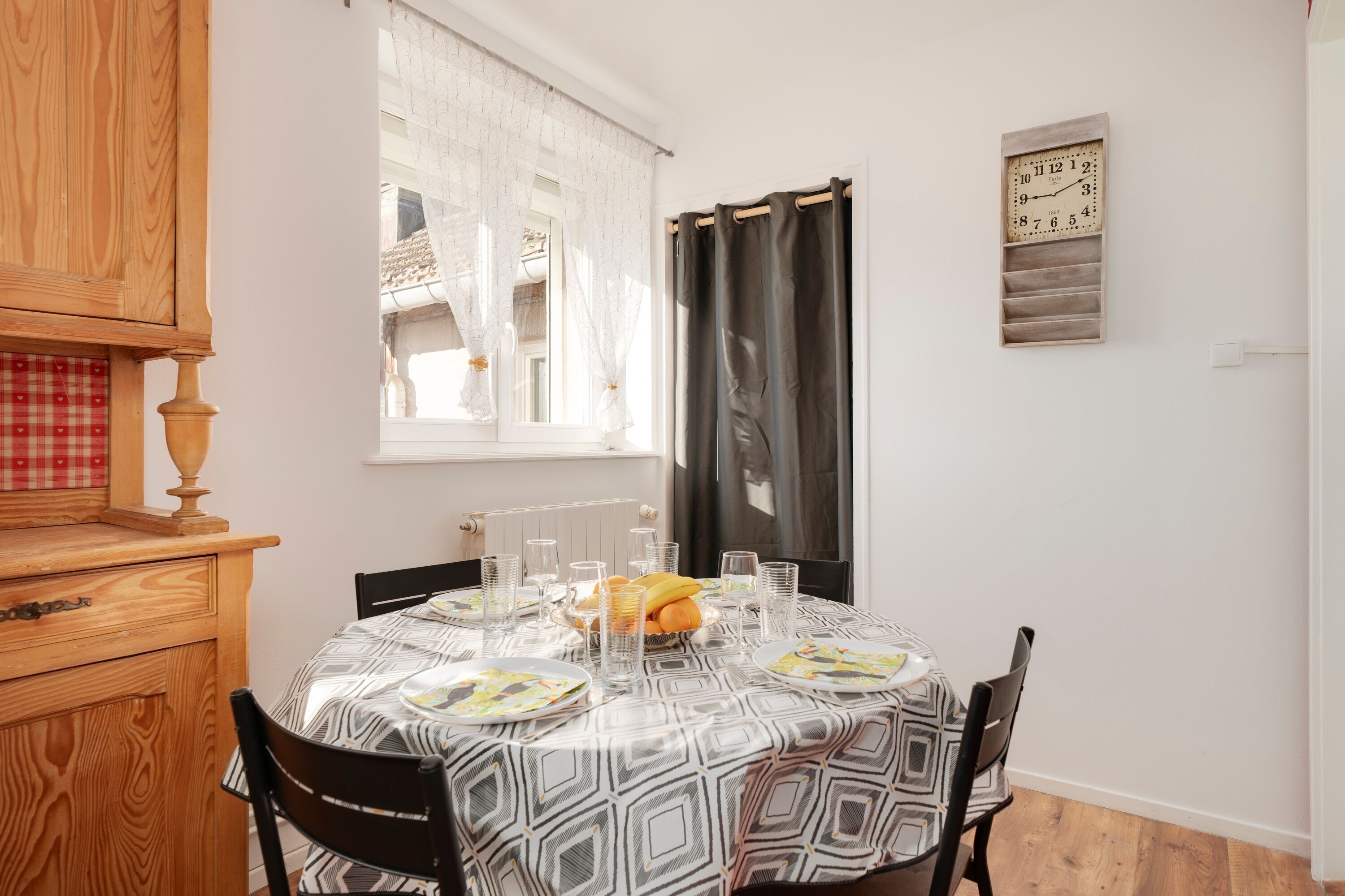 A dining area with a round table set for four, a wooden cabinet, a window with curtains, and a wall-mounted clock.