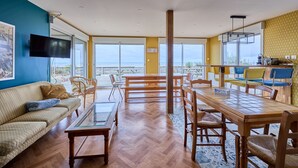 Un vaste espace de vie comprenant une grande table à manger en bois, des sièges confortables, un téléviseur à écran plat et de grandes fenêtres offrant une vue sur la mer.