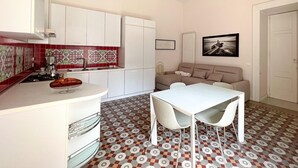 A compact kitchen with white cabinets, a red and white tiled backsplash, a gas stove, and a white countertop. There is a dining area with a white table and two chairs. - YourHome - Chiara's Apartment Sorrento 2 (Piano di Sorrento)