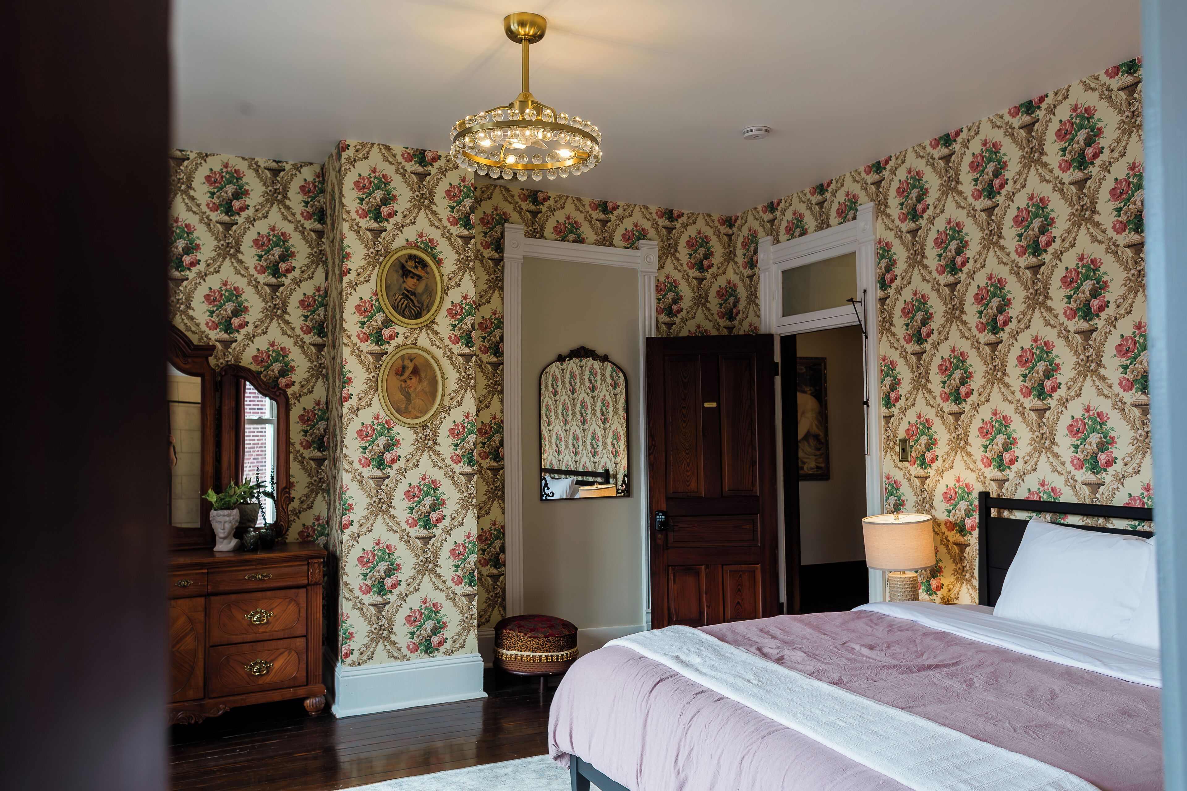 A bedroom with floral wallpaper, a wooden dresser, a round ottoman, a bed with a white and pink bedspread, and a chandelier.