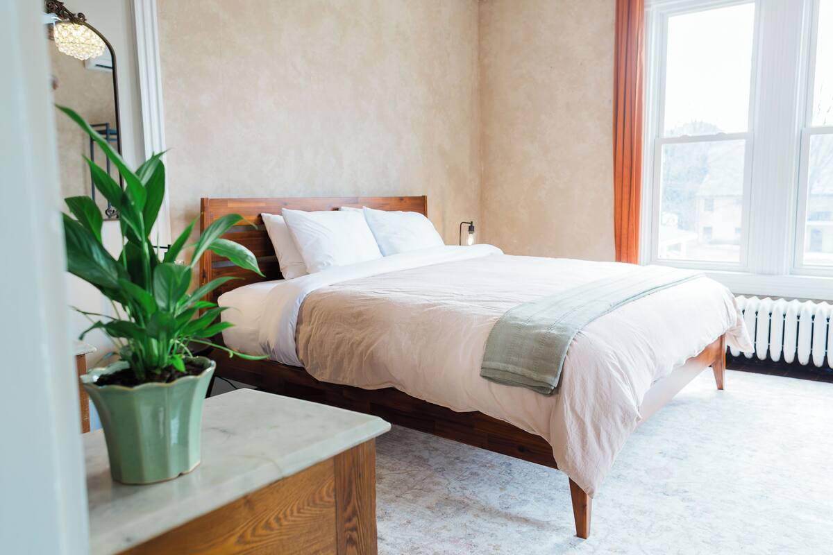 A neatly made bed with a wooden headboard, a potted plant on a bedside table, and a window with curtains.