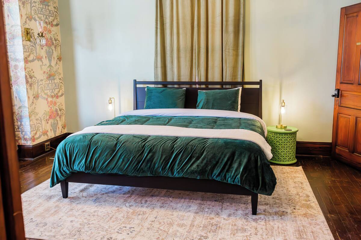 A bed with a dark headboard, a green and white bedspread, and two pillows. There is a green side table with a lamp, and a wooden door is visible to the right.
