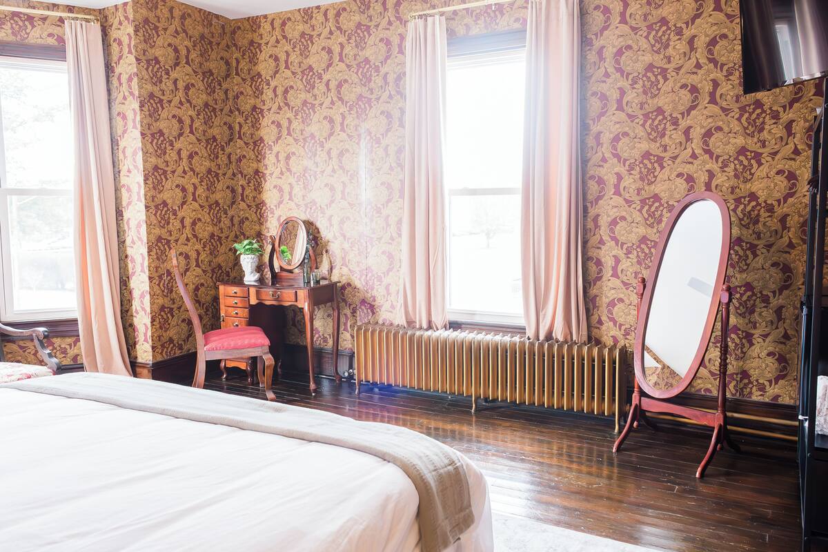 A hotel room with a large bed, a wooden dresser, a chair, a mirror, and a radiator.