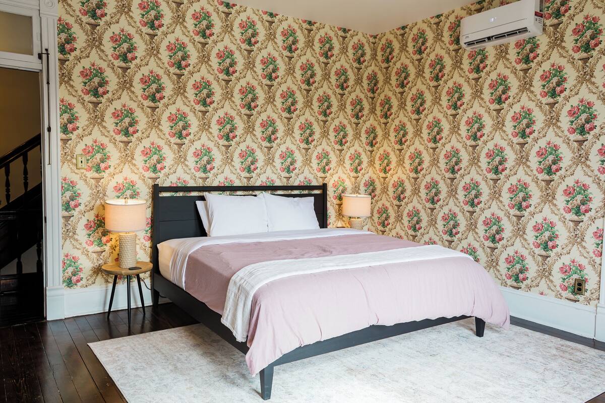 A bedroom with a bed, two bedside lamps, a patterned wallpaper, and a staircase.