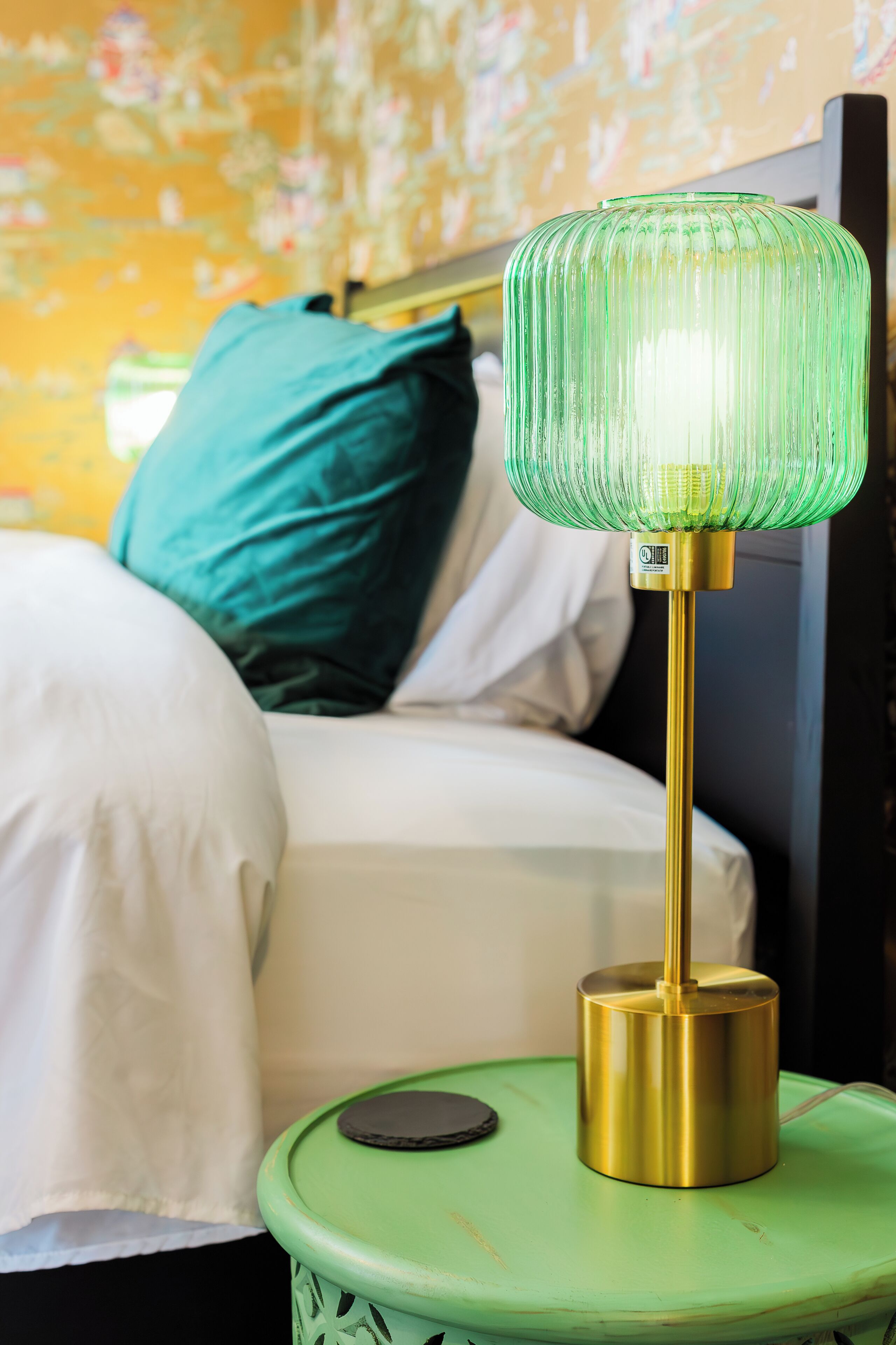 Uma cama com lençóis brancos, um criado-mudo verde e uma luminária com base dourada.