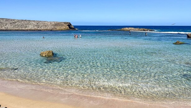 A clear beach with shallow water, rocks, and a distant cliff.