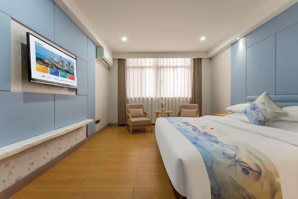 A hotel room with a bed, a TV mounted on the wall, a small table, two chairs, and a window with curtains.