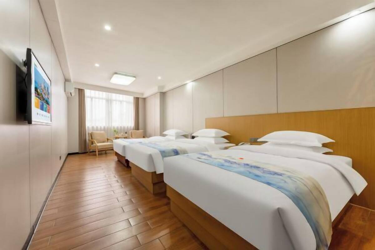Ein Hotelzimmer mit zwei Betten, Holzboden, einem an der Wand befestigten Fernseher und einem Fenster mit Vorhängen.