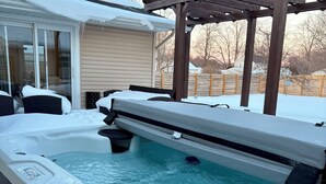 Un jacuzzi con una terraza de madera y una pérgola en un patio trasero nevado.