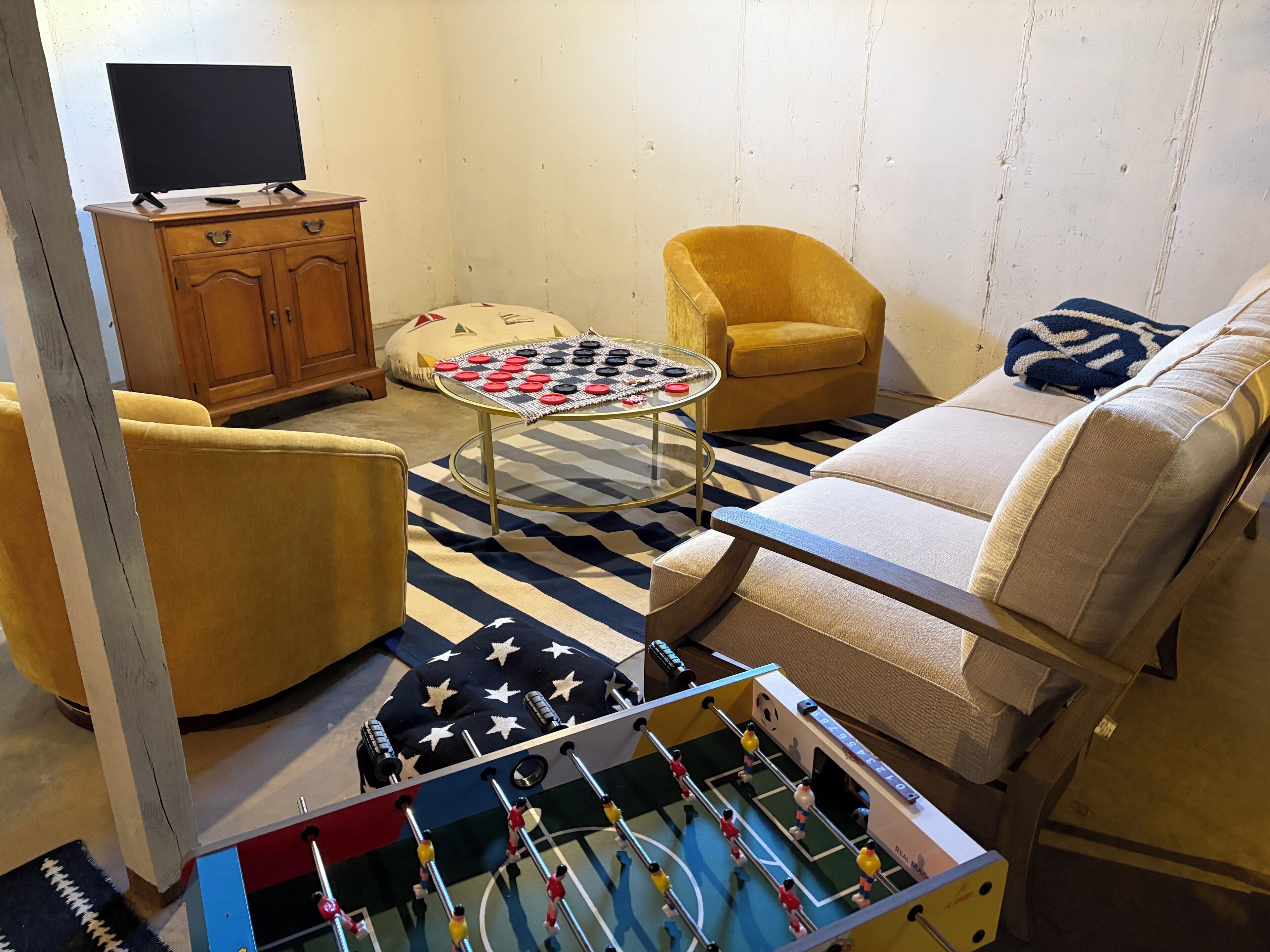 Una sala de estar con mesa de fútbol, sofá, sillón amarillo, televisor sobre un mueble de madera y alfombra estampada.