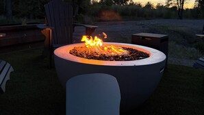 Ein Feuerkorb mit Flammen, umgeben von Gartenmöbeln und Bäumen.