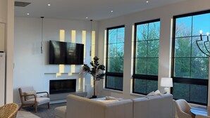 Ein modernes Wohnzimmer mit großem Fernseher, Kamin, Sofa und großen Fenstern.
