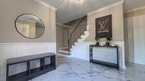 A hallway with a marble floor, a round mirror, a Louis Vuitton sign, a staircase, and a console table with plants.