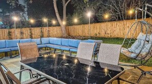 Uma área externa com mobiliário de jardim, incluindo uma mesa com tampo de vidro, sofás com almofadas azuis e uma cadeira suspensa.