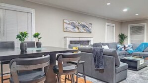 A modern living room with a grey sofa, a coffee table, and a fireplace. There are two chairs and a dining table with potted plants.