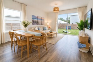 A spacious living area with a dining table, wooden chairs, a sofa, and a TV mounted on the wall. - Family Retreat - Arcade, Fire Pit, Game Room, King Bed - 2 mi Downtown! (St. Augustine)