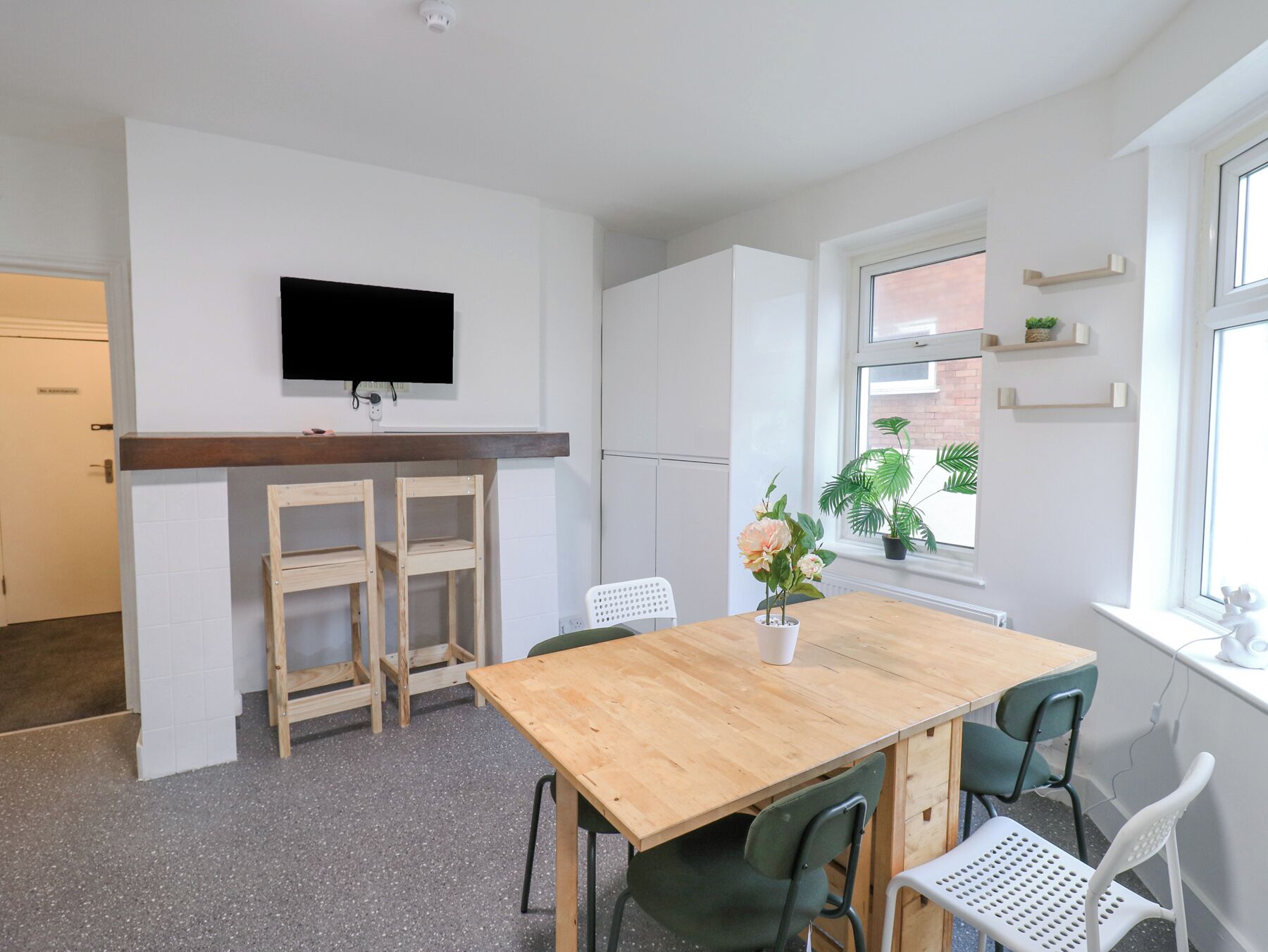 A compact kitchen with a wooden table and chairs, a flat-screen TV mounted above the counter, and a window with a view of the outside.