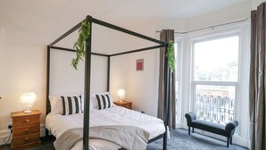 Una cama con dosel con ropa de cama blanca, una mesita de noche de madera con lámpara, un banco junto a la ventana y vistas al exterior.