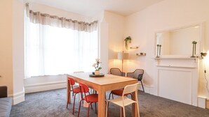 Un comedor con una mesa de madera, cuatro sillas y un florero. Hay una ventana con cortinas translúcidas y una lámpara sobre una mesita.