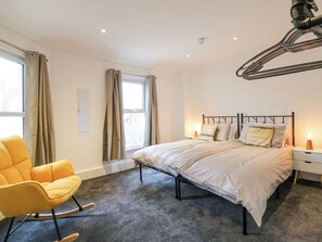 A modern bedroom with a bed, a rocking chair, a window with curtains, and a wall-mounted lamp. - The Wiltons (Bexhill-on-Sea)