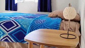 Quarto com uma cama, uma mesa redonda de madeira, um ventilador e uma janela com cortinas.