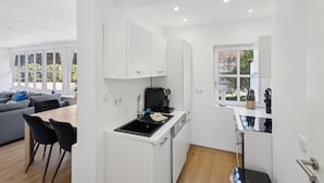 Una cocina moderna con armarios blancos, una cafetera y un lavabo. Hay una zona de comedor con una mesa y sillas.