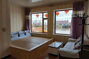 A room with a bed, a small table, and a sofa. There are windows with curtains and a view outside.