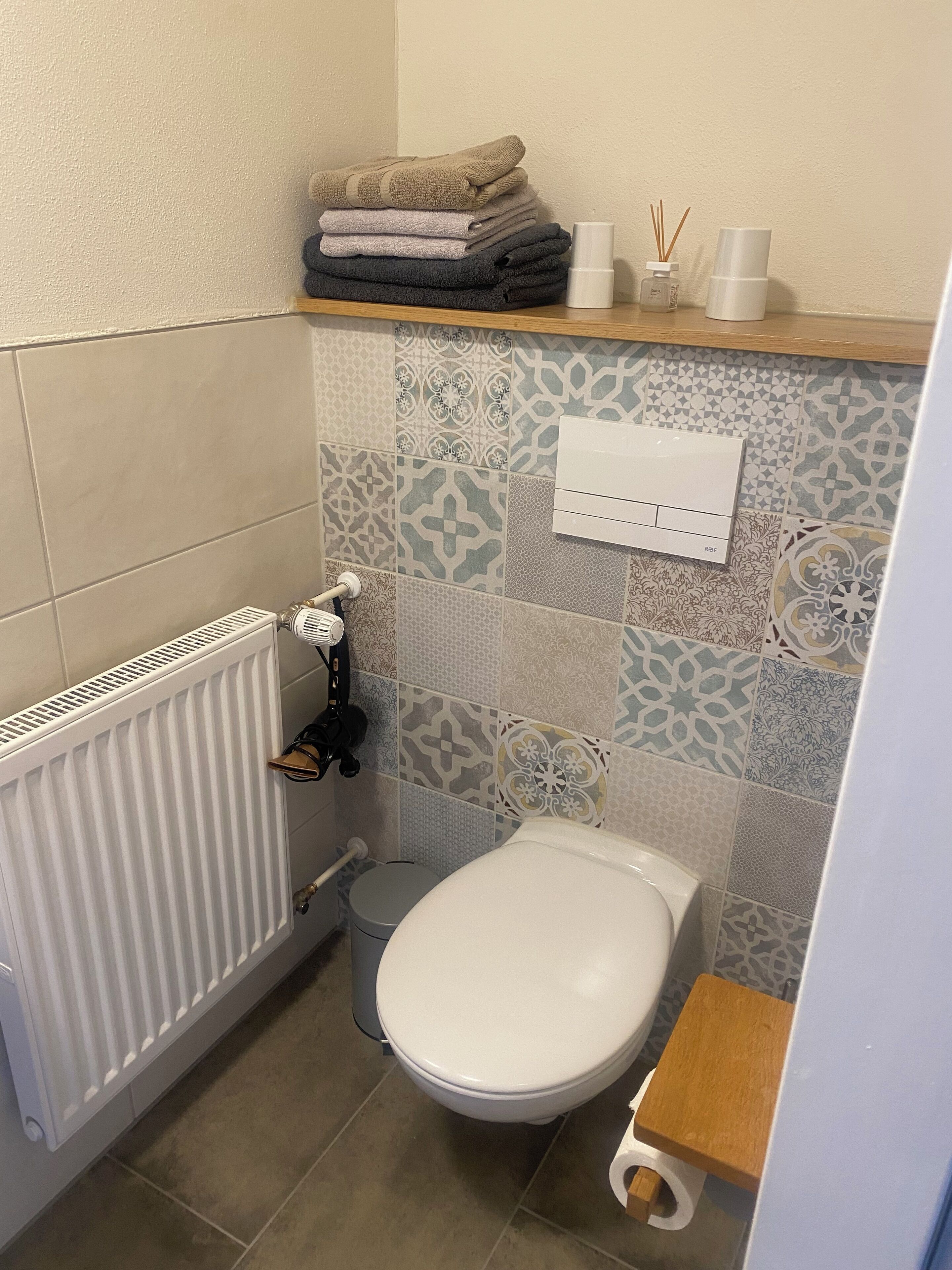Un baño pequeño con inodoro, un radiador, azulejos estampados, un estante con toallas y un dispensador de papel higiénico.