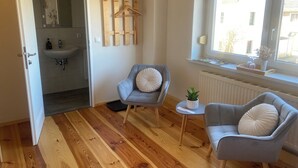 Una habitación con piso de madera, un sillón gris con un cojín blanco, una mesita con una planta y una ventana con vista a edificios.