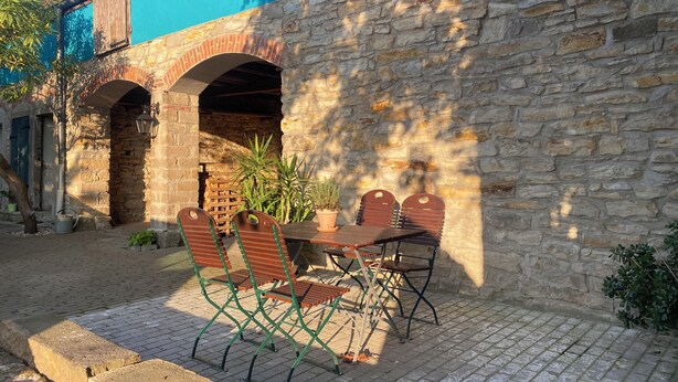Un patio con pared de piedra, una mesa de madera y sillas de metal verde.