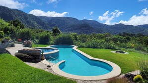 Una piscina de diseño curvo, rodeada de césped y plantas cuidadas, con montañas y cielo despejado al fondo.