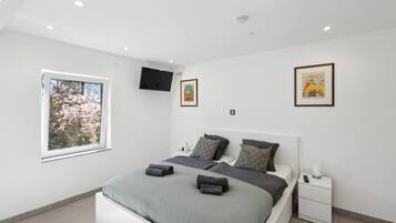 Ein modernes Schlafzimmer mit einem großen Bett, einem Nachttisch, einem an der Wand befestigten Fernseher und einem Fenster mit Blick auf Bäume.