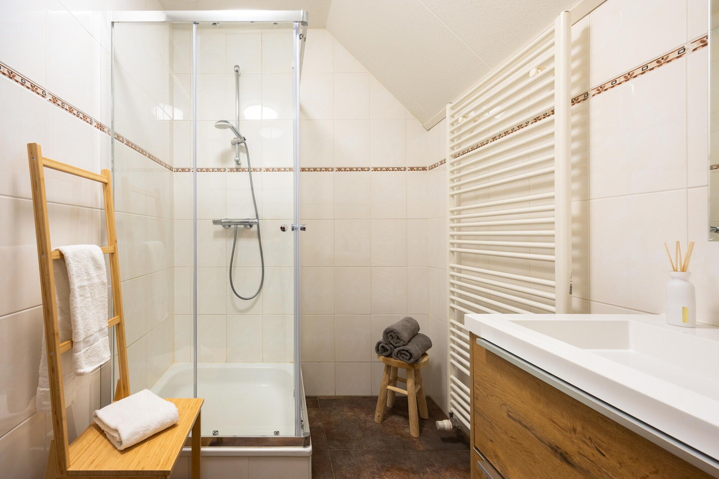 A bathroom with a glass-enclosed shower, a wooden towel rack, a wooden stool with towels, a white sink, and a radiator.
