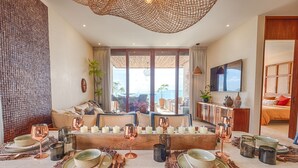 Uma área de refeições com vista para o mar, um sofá e uma TV ao fundo.