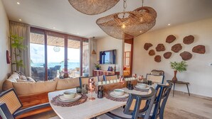 Uma sala de jantar com uma mesa grande posta para seis pessoas, rodeada por cadeiras com encostos trançados. Há velas sobre a mesa, e uma televisão está montada na parede.