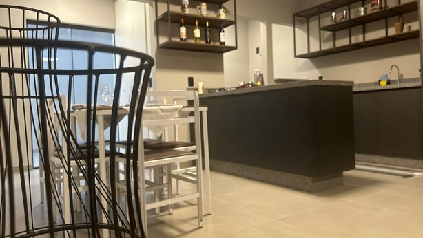 A modern kitchen with a central island, white chairs, and built-in shelves with kitchenware. - GEMINADO CASA 4 ALTO PADRÃO 3 QUARTOS (Joinville)