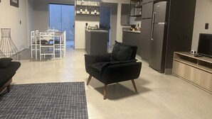 A modern living room with a black armchair, a dining area with a white table and chairs, a kitchen with dark cabinets and a refrigerator, and a staircase leading to an upper level. - GEMINADO CASA 4 ALTO PADRÃO 3 QUARTOS (Joinville)