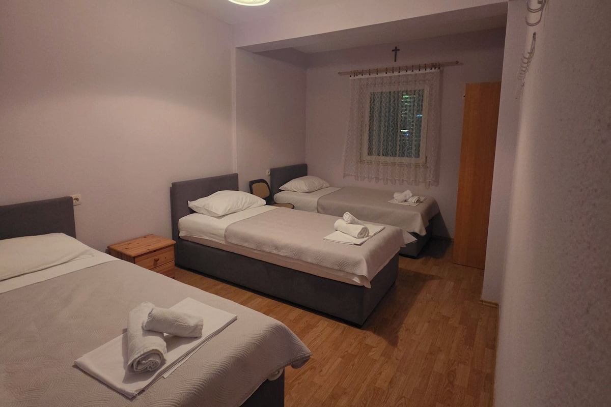 A hotel room with two beds, a small wooden nightstand, a window with curtains, and a wall-mounted light fixture.
