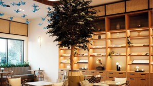Een modern interieur met een centraal zitelement, houten planken met boeken en decoratieve objecten, en een grote boom in het midden.