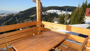 Een houten terras met een tafel en banken, met uitzicht op een besneeuwd landschap met bomen en een verre brug.